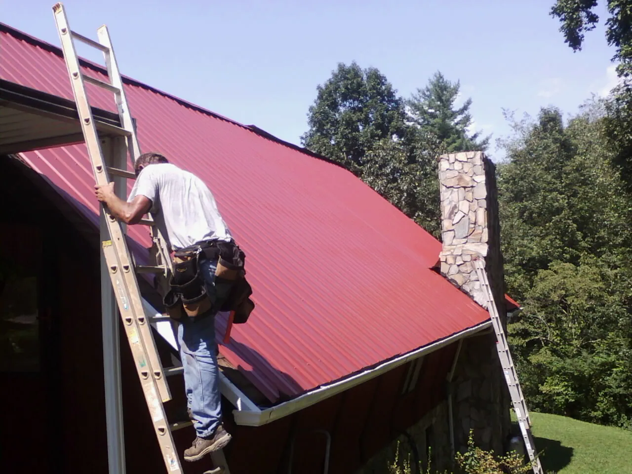 Roofer on a ladder beside a red metal roof and stone chimney — Soffit & Fascia Repair by Roofing Brothers in Richboro